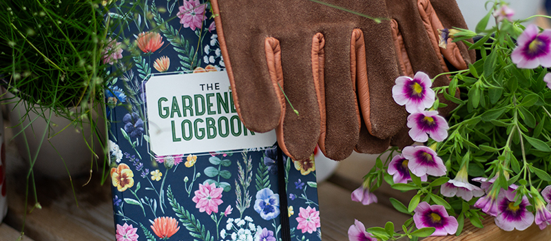 Gardening gloves resting on a floral garden logbook next to blooming flowers and potted plants